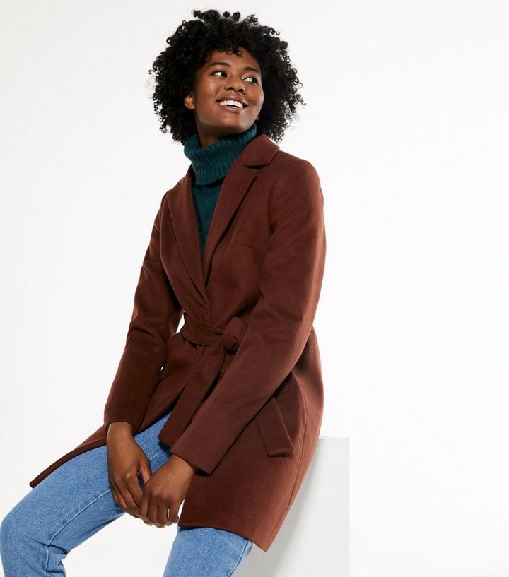 Vegan coat in a wine red colour with belt worn by a woman who is also wearing bluejeans