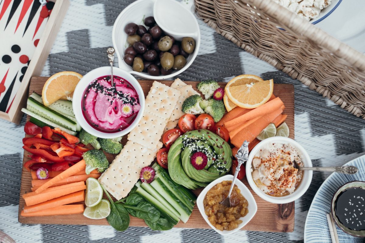 Picnic Spread with Vegetables, Crackers, Dips and Fruit on Grey and White Blanket.