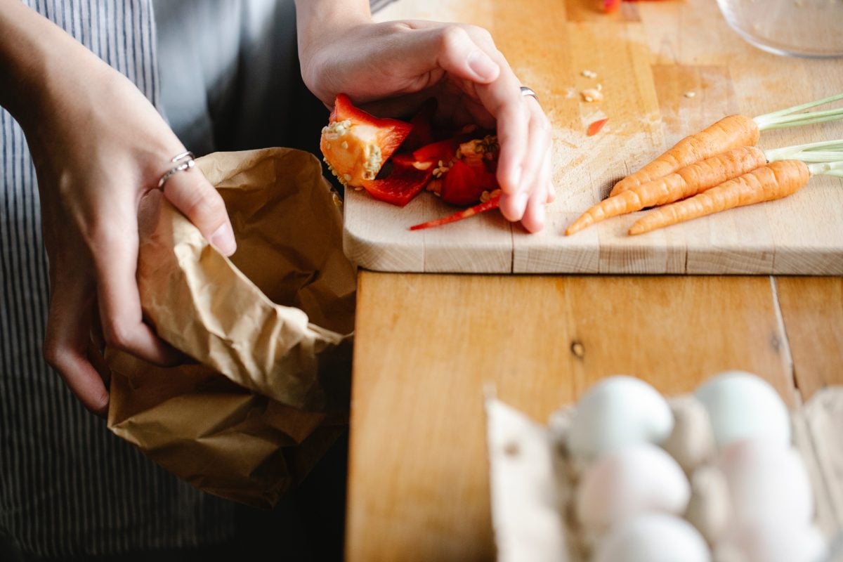 A close up of a hand pushing food scraps such as peppers of a wooden cutting board into a paper bag