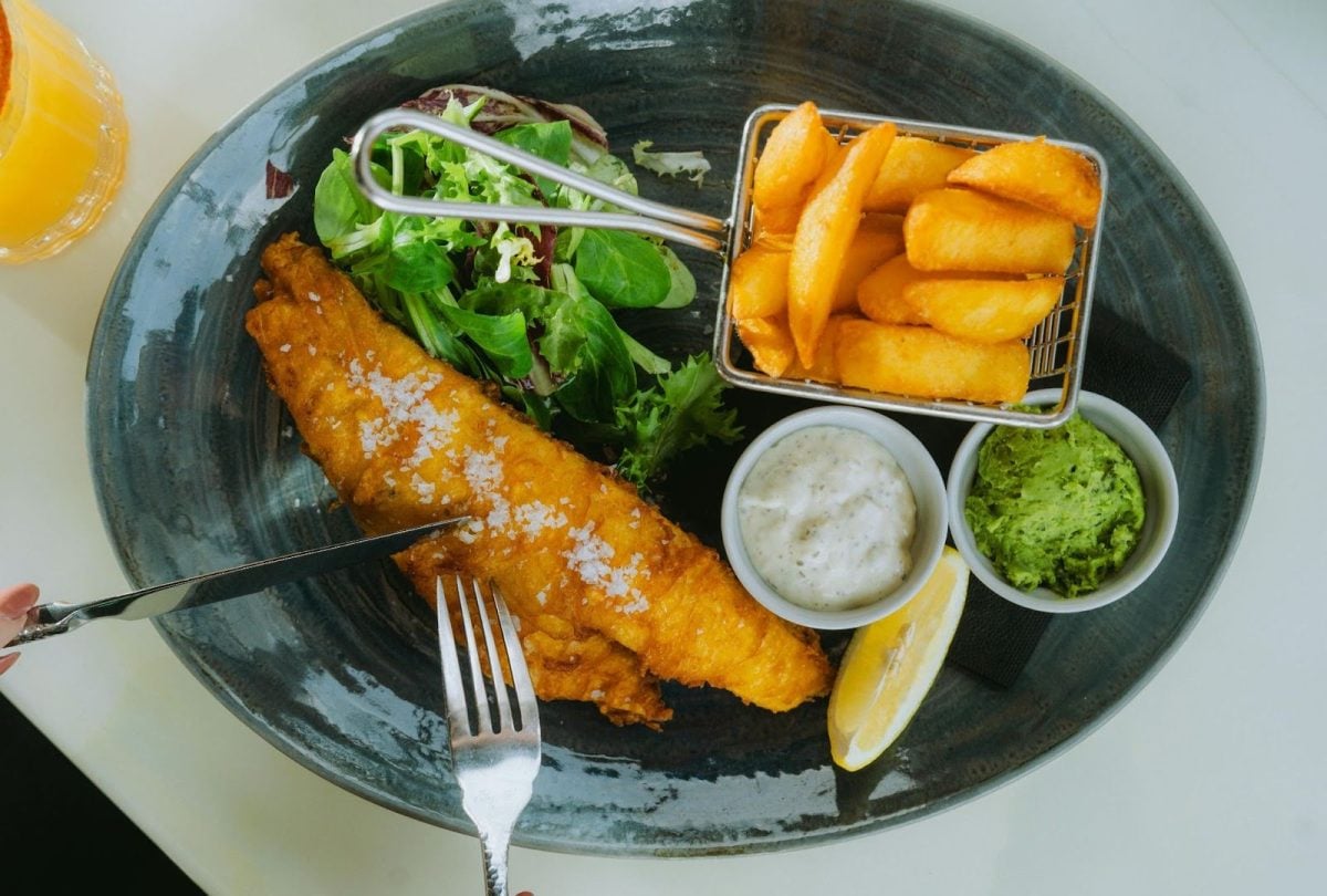 Person cutting into a plate of fish and chips, with salad and sauces